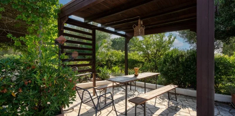 An outdoor dining area sits beneath a wooden pergola, surrounded by greenery.