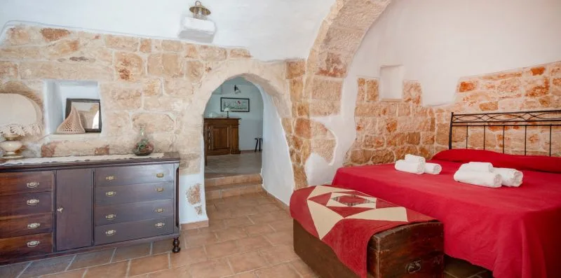 A bedroom with a red bedspread and a stone archway leading to another room.