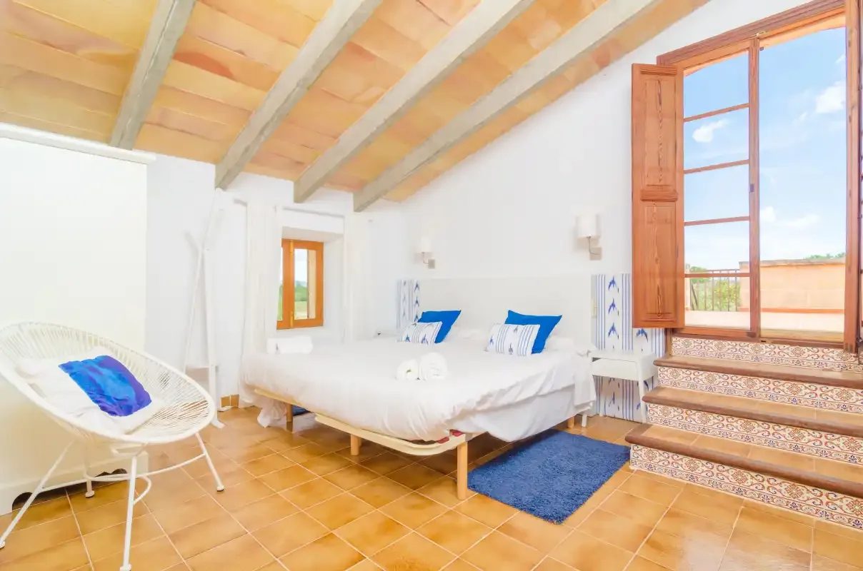A bright bedroom has white bedding, a woven lounge seat, and a tall open window.