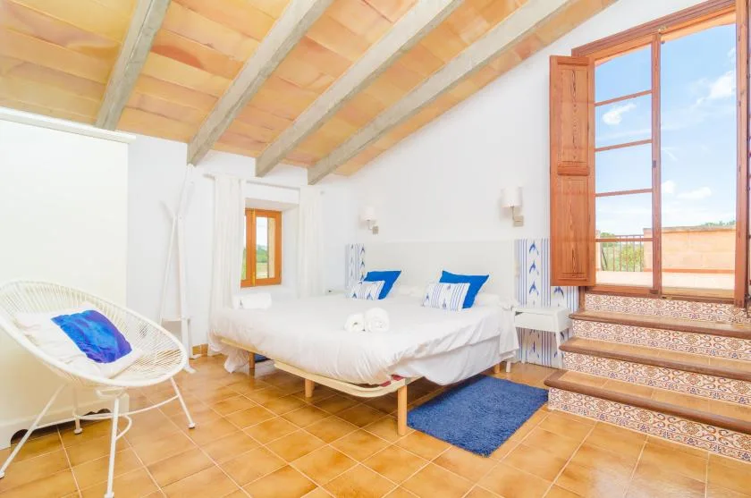 A bright bedroom has a bed, a woven chair, and an open window.