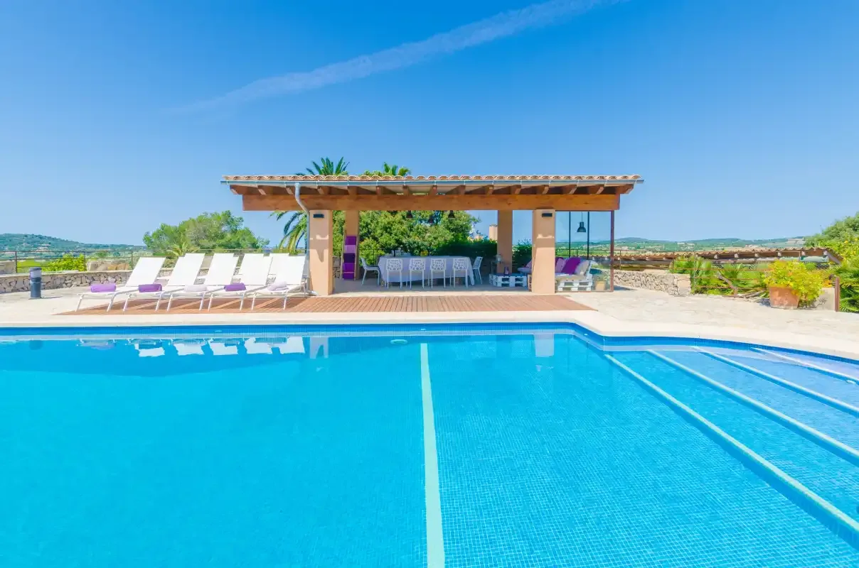 A swimming pool fills the foreground, with a shaded seating area and loungers beyond.