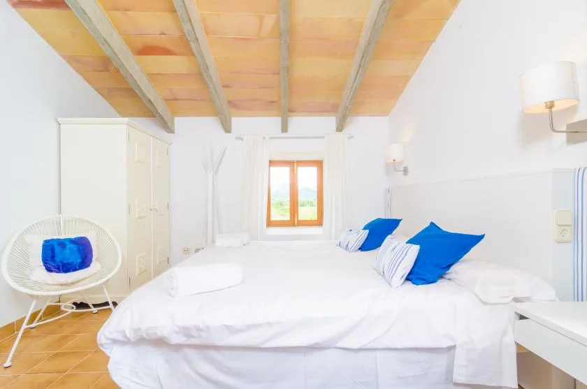 A bright bedroom with white bedding, blue pillows, and exposed ceiling beams.
