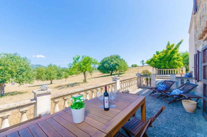 A wooden dining table sits on a stone terrace overlooking a sunny landscape.