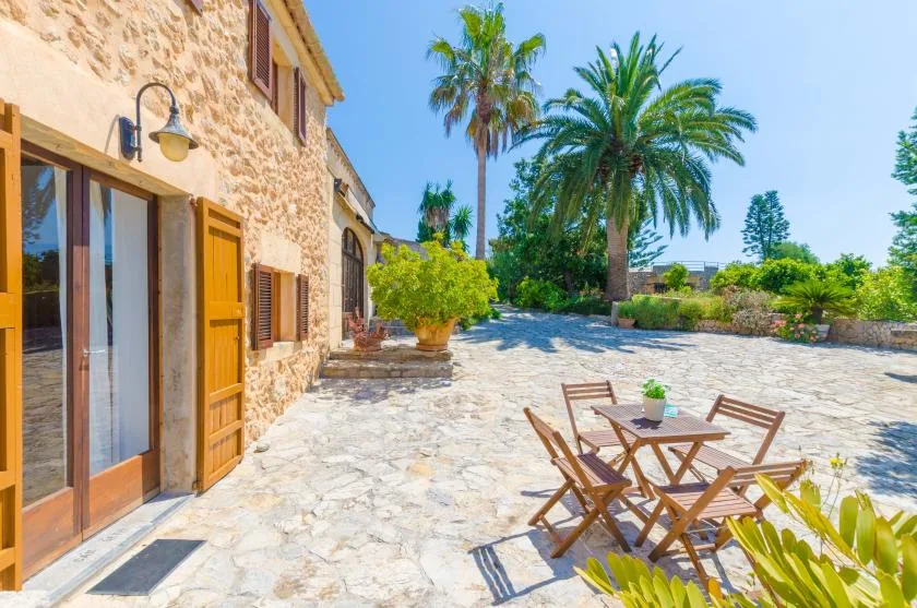 A stone building opens onto a sunny stone patio with outdoor seating and palm trees.