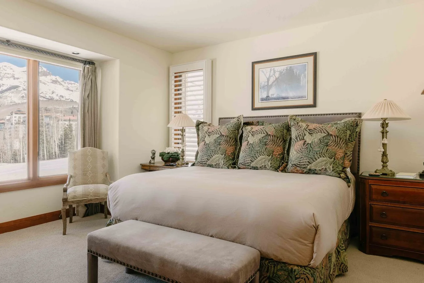 A bedroom with a bed, a cushioned bench, and a window overlooking snowy mountains.