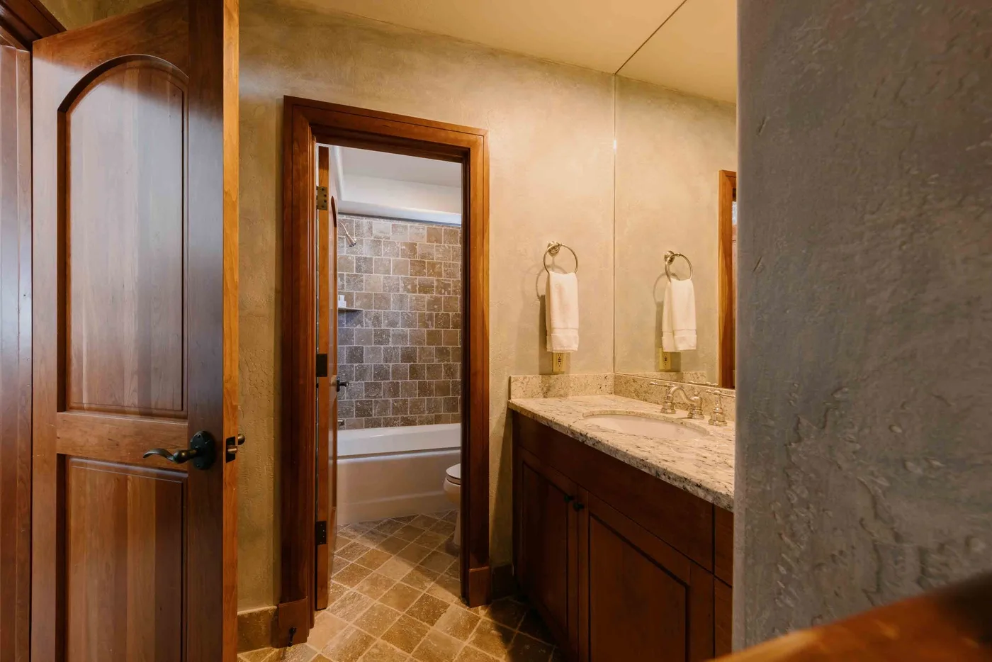 A bathroom vanity with a mirror beside an open doorway to a tiled bathtub.