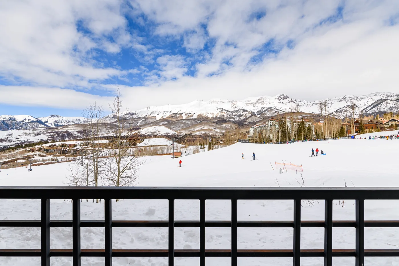 A snowy ski slope stretches toward mountain peaks beneath a partly cloudy sky.