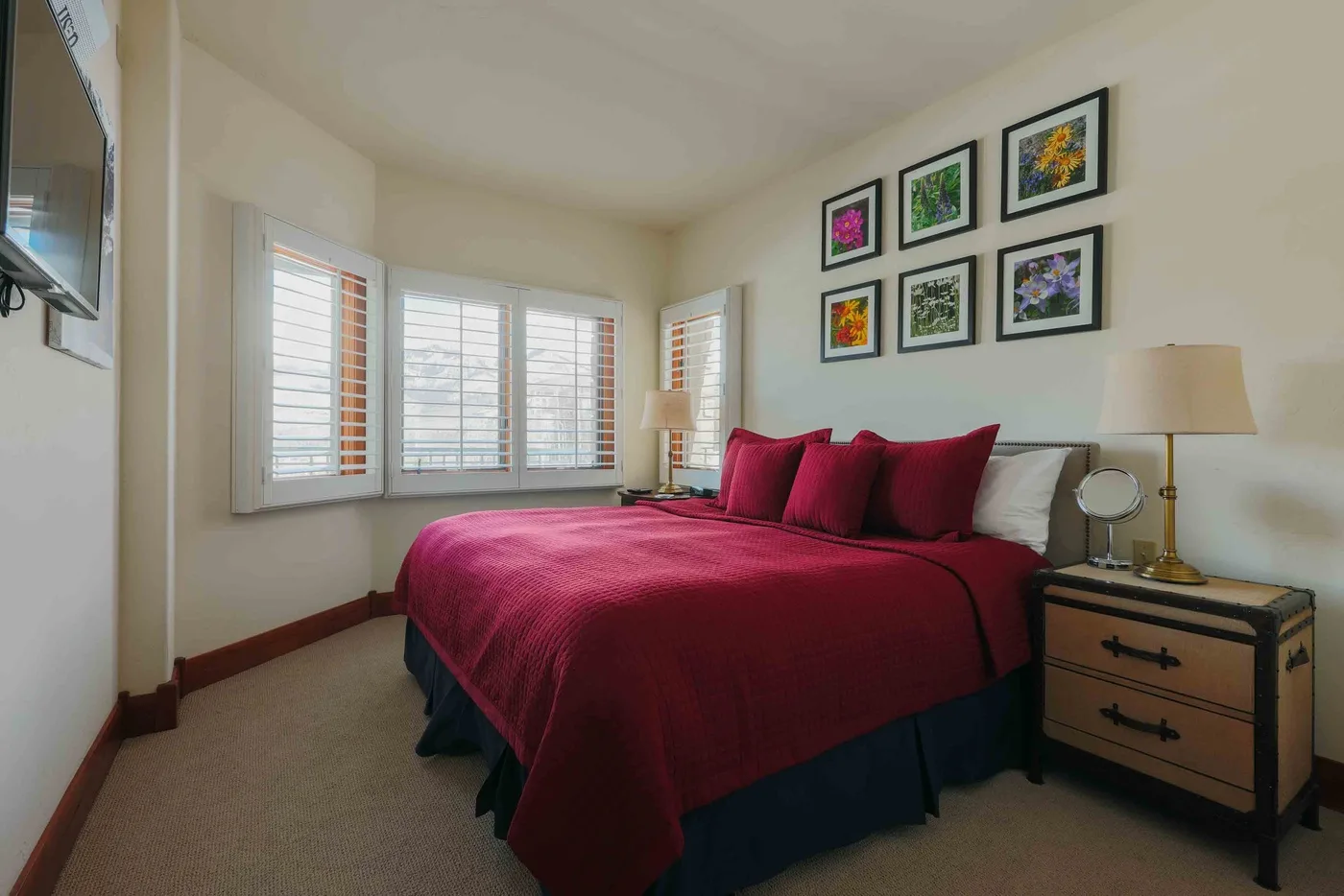 A bedroom with a burgundy bedspread, windows, and framed artwork.