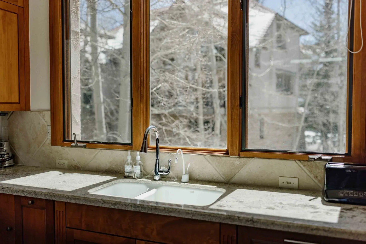 Kitchen sink and counter beneath large windows overlooking trees and a neighboring building.
