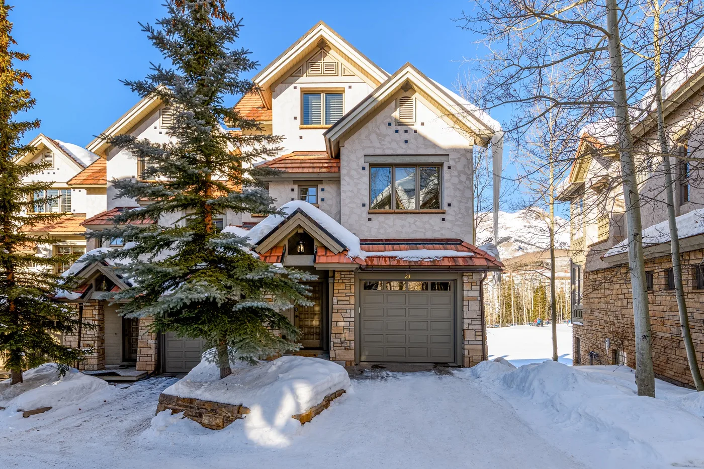 A multi-story house stands beside a snowy driveway, with trees around it.