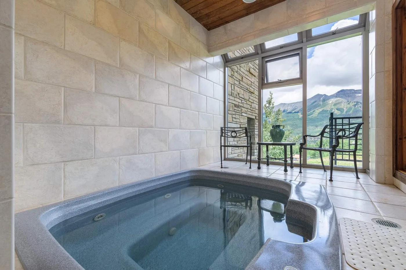 Una piscina de spa interior con azulejos está junto a ventanales con vistas a montañas verdes.