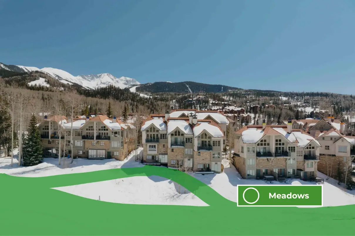Snow-covered mountain lodges sit below a wooded hillside under a clear sky.