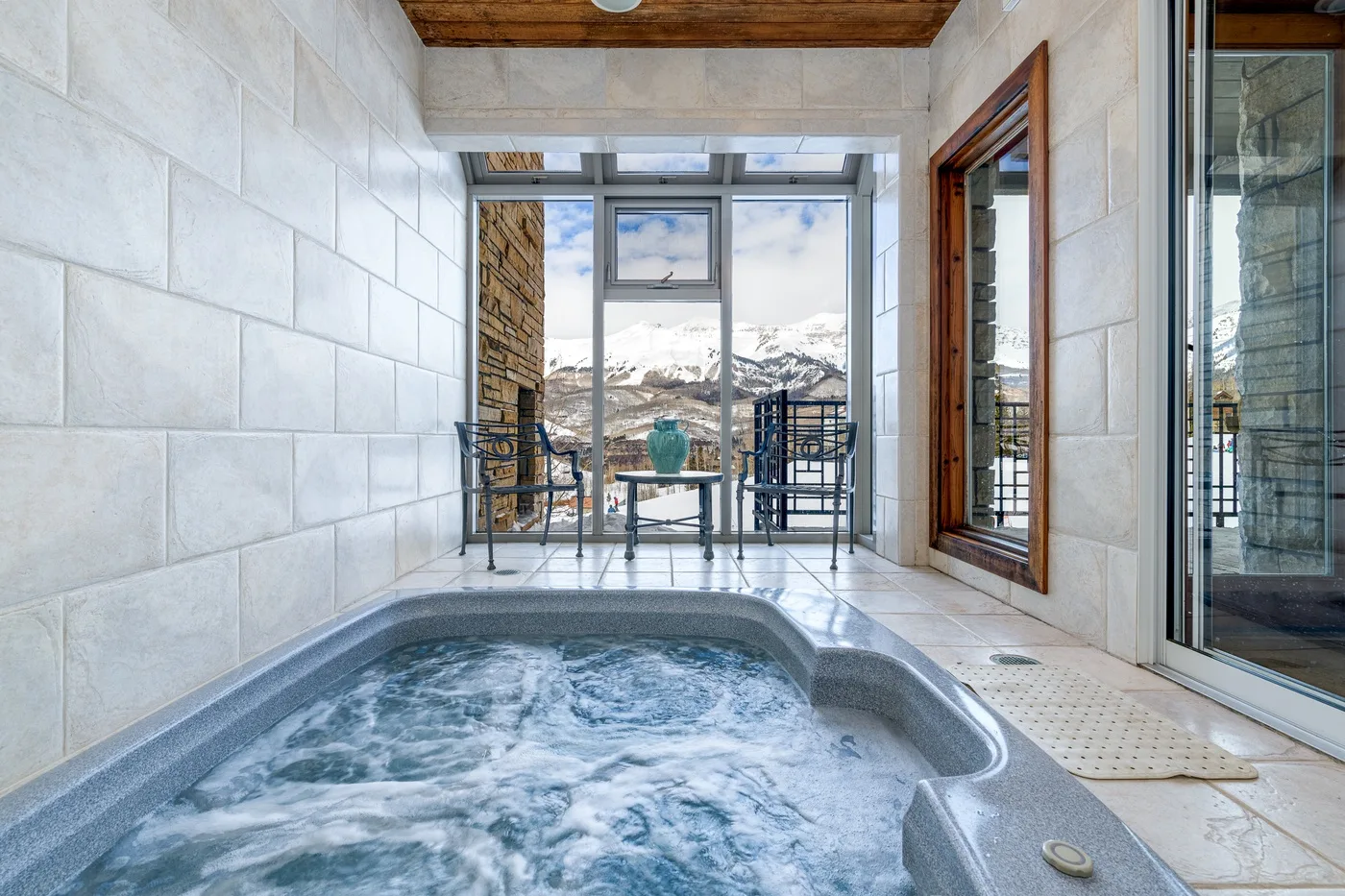 An indoor hot tub sits beside windows overlooking a snowy mountain landscape.