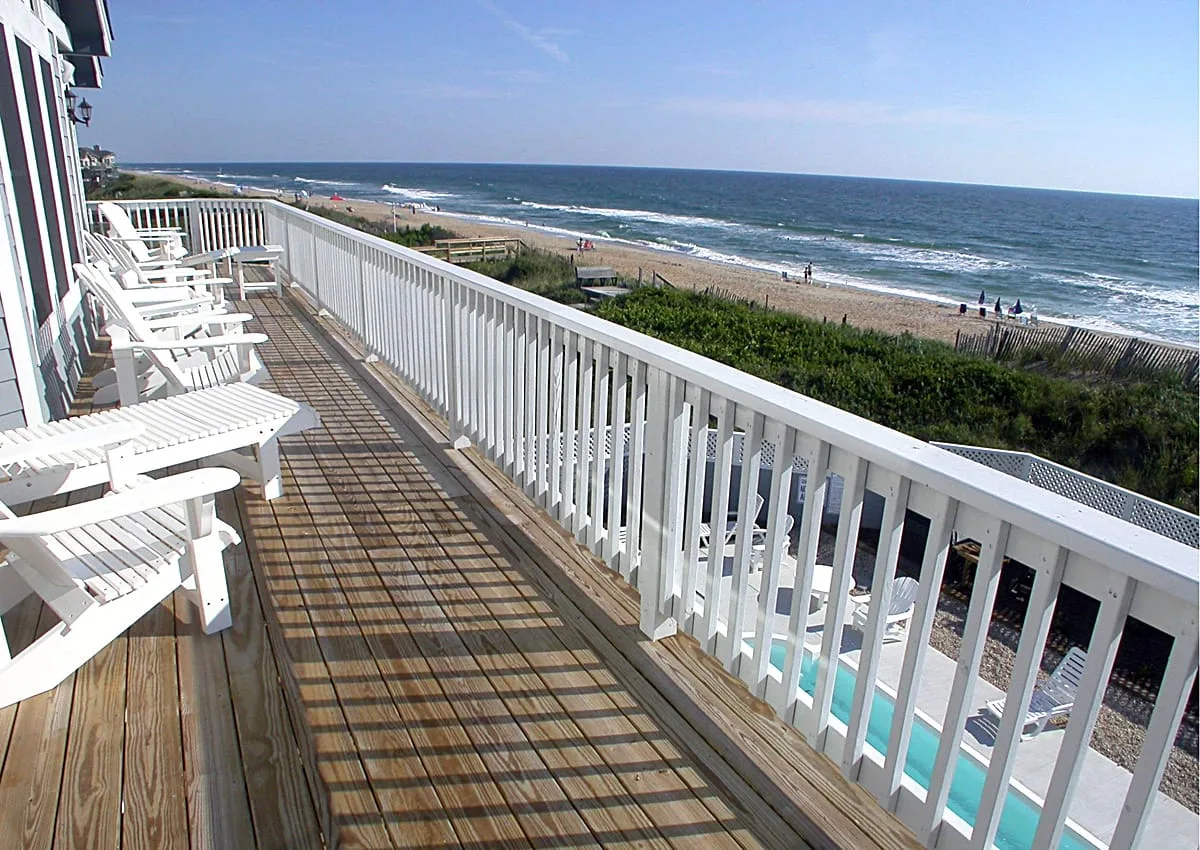 Una terraza de madera con asientos blancos da al mar y la playa.