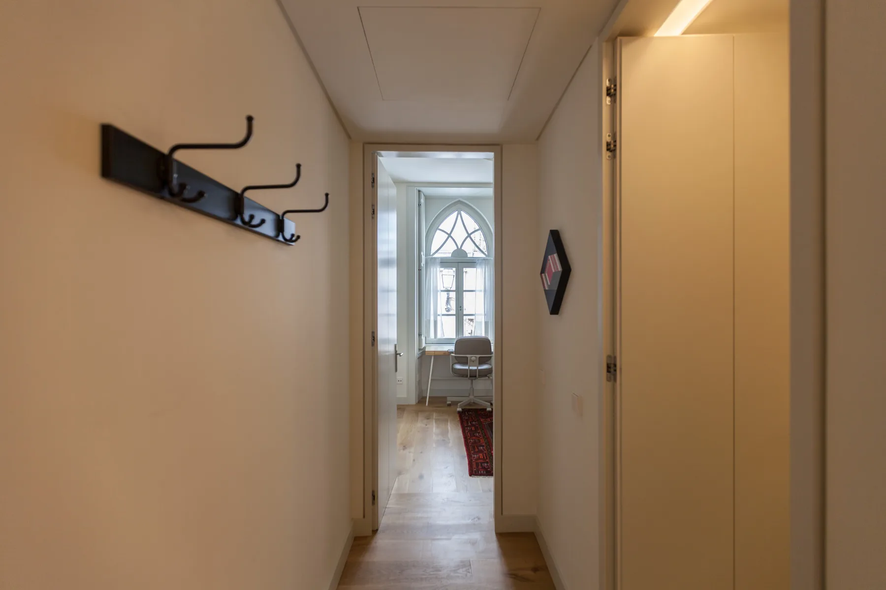 A narrow hallway with wall hooks leads toward a bright room.