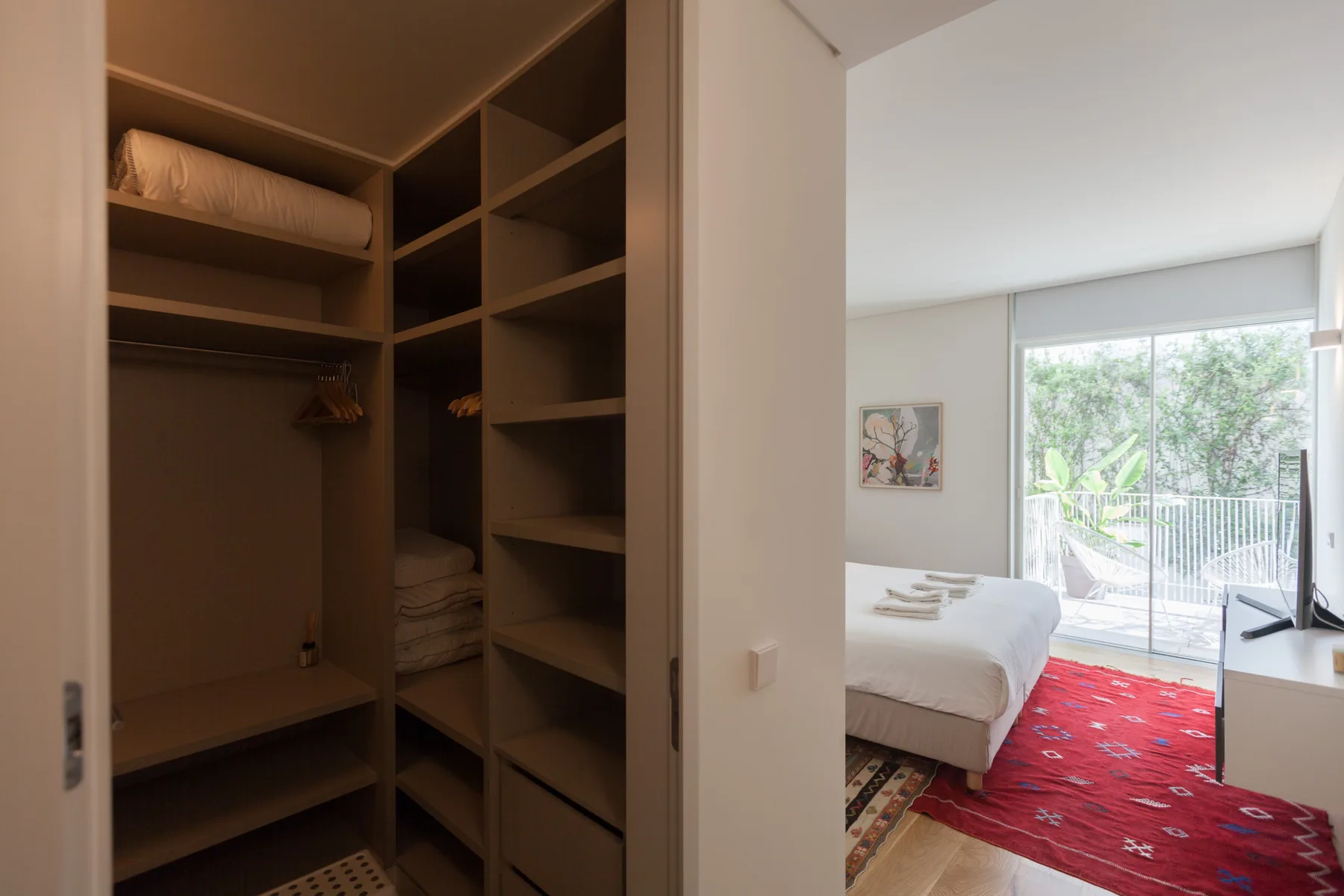 An open wardrobe overlooks a bedroom with a bed and red rug.