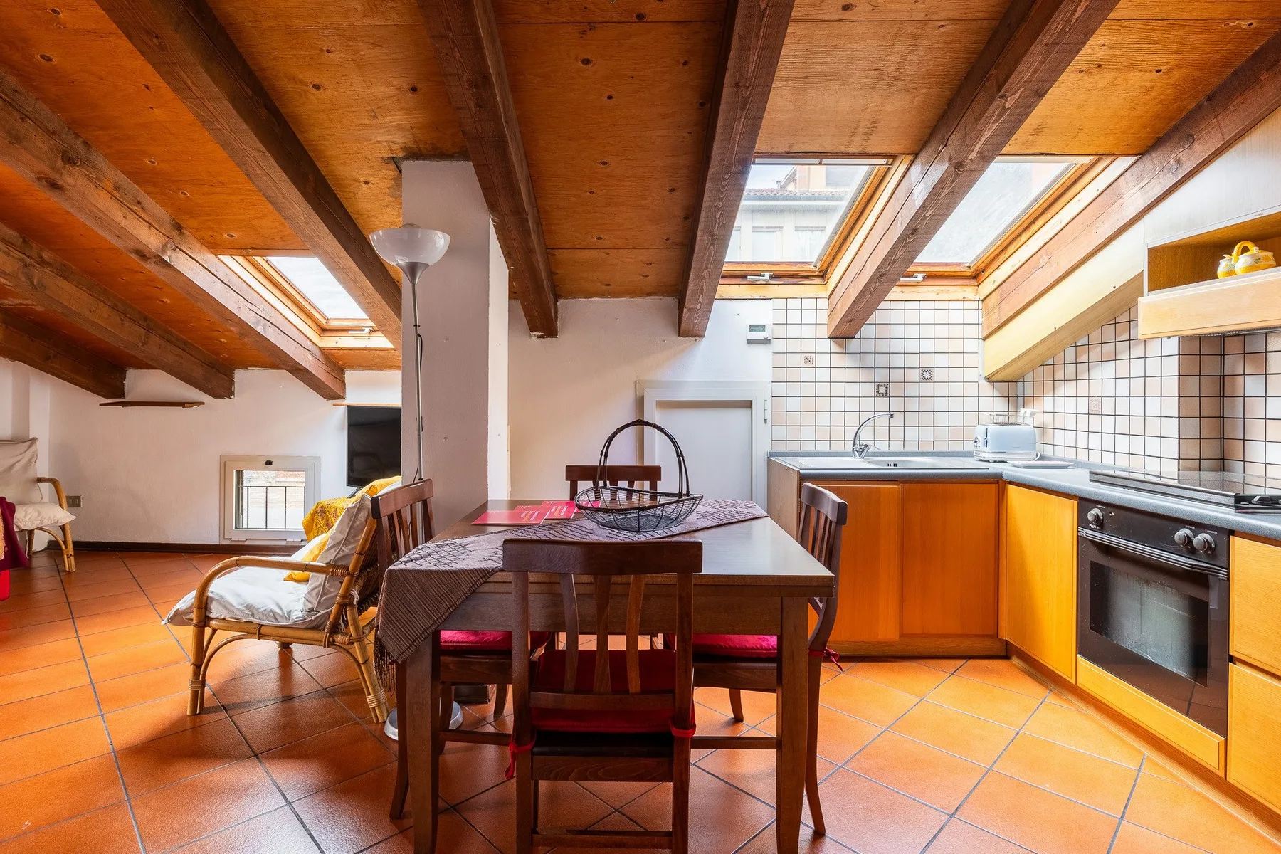 Zona de comedor y cocina con una mesa de madera bajo vigas vistas y tragaluces.
