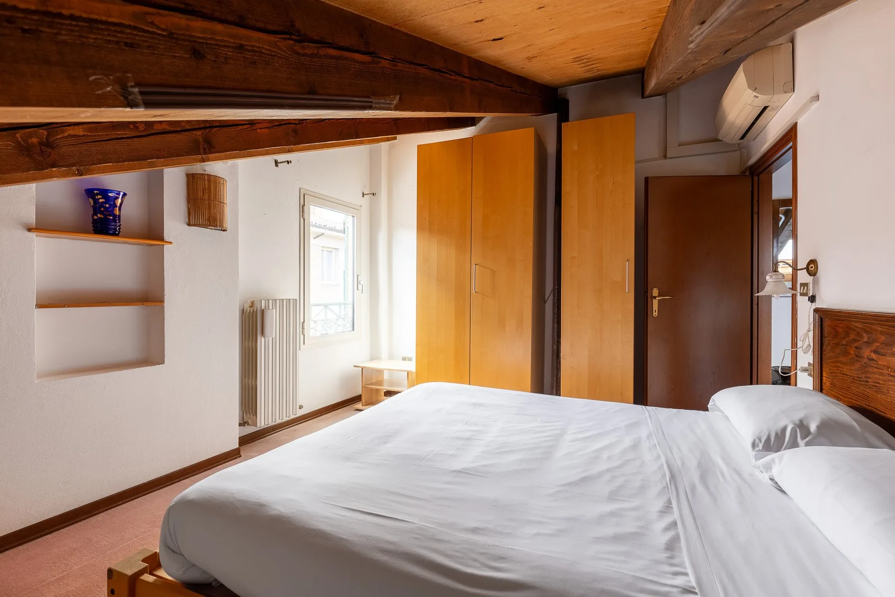 Un dormitorio luminoso con una cama, vigas de madera y estanterías empotradas.