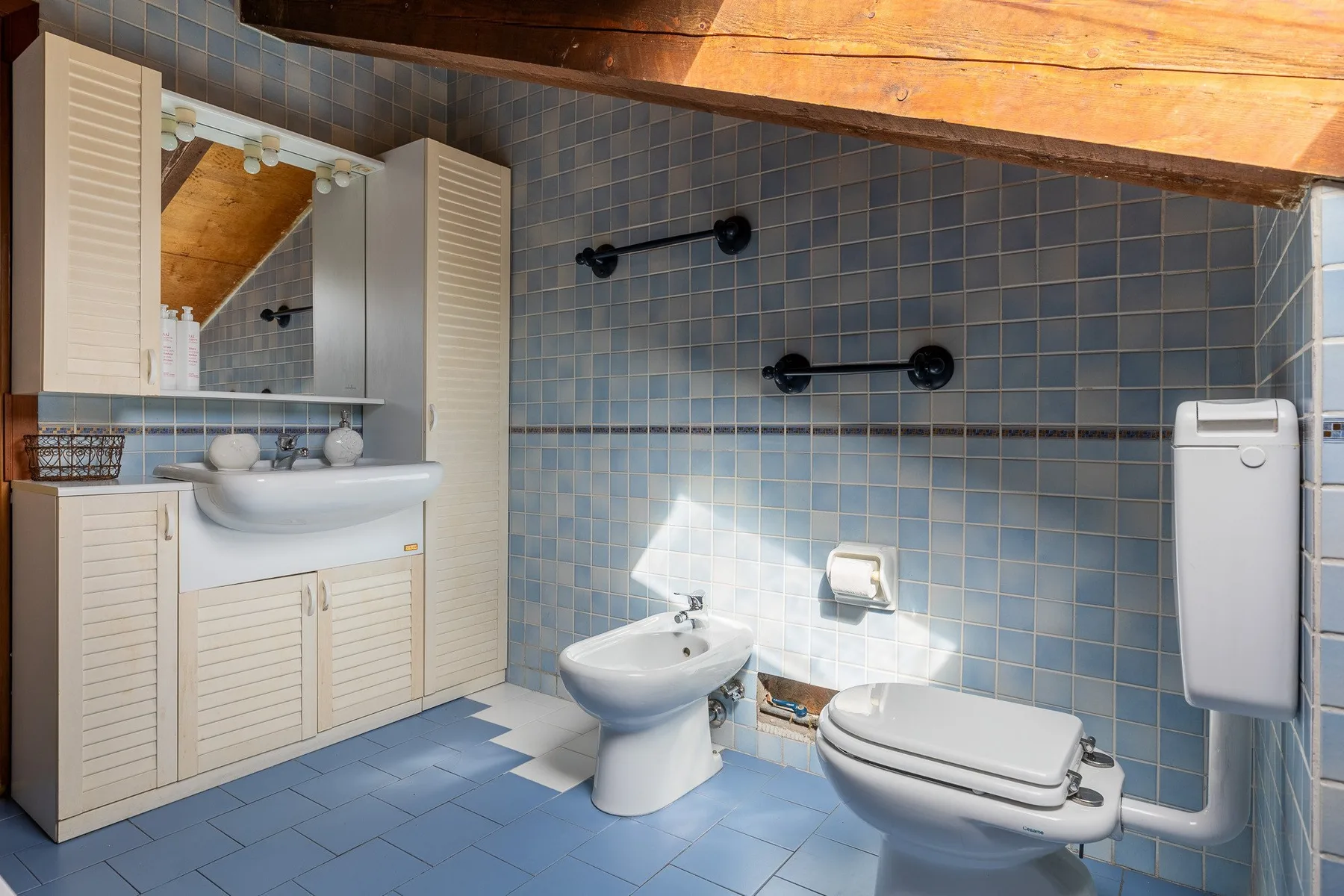 Un baño con azulejos azules, lavabo, bidé e inodoro bajo un techo inclinado de madera.