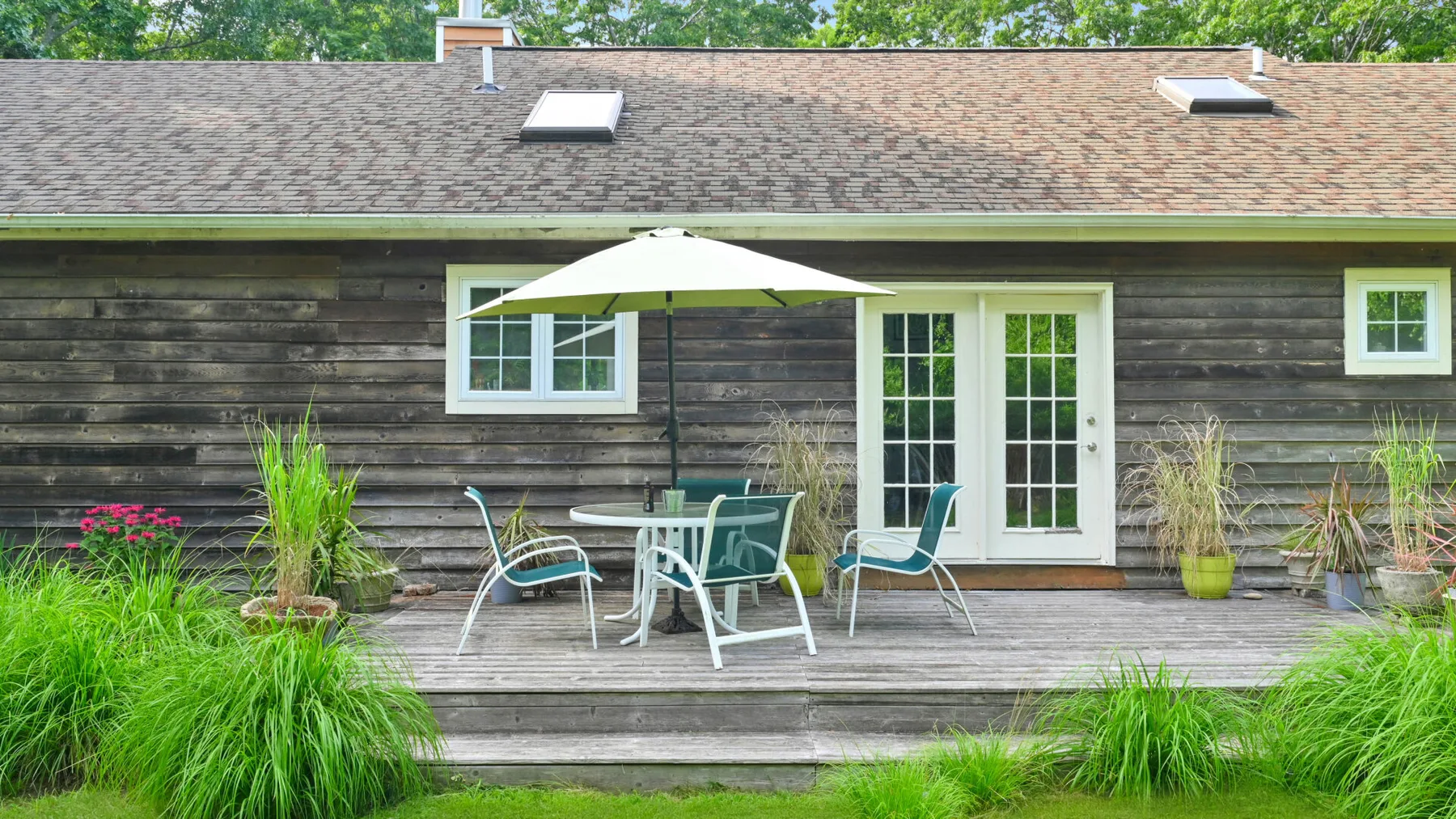 A wooden house opens onto a deck with a table, chairs, and umbrella.