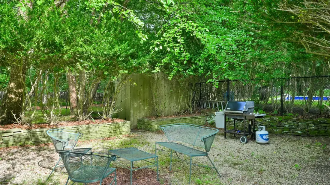 A shaded outdoor seating area with metal chairs and a small table beneath leafy trees.