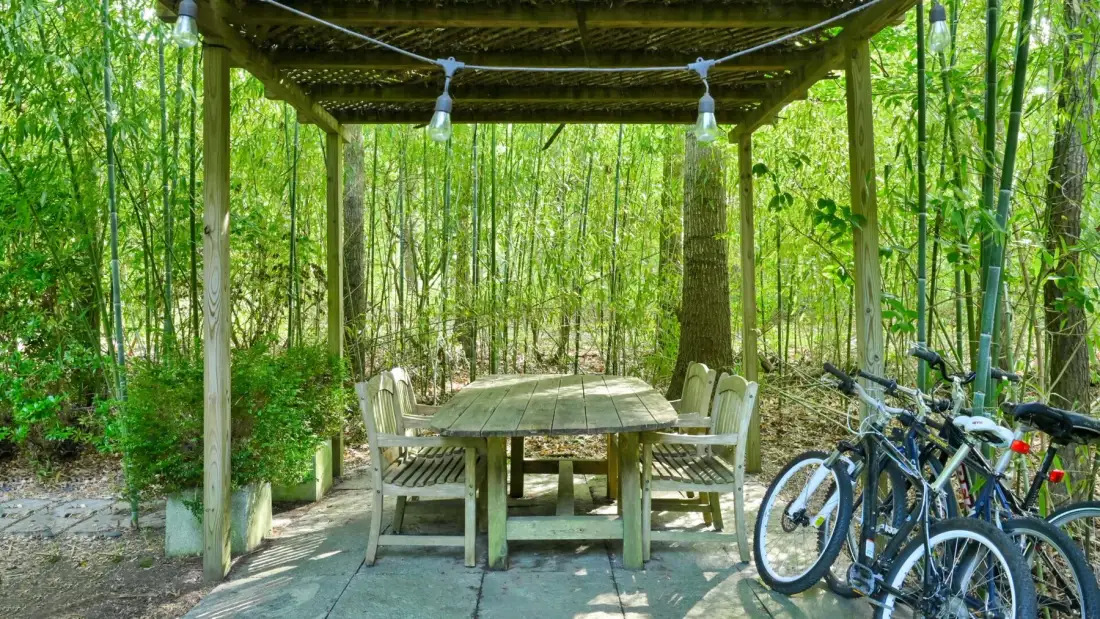 An outdoor dining area beneath a wooden pergola, surrounded by greenery.