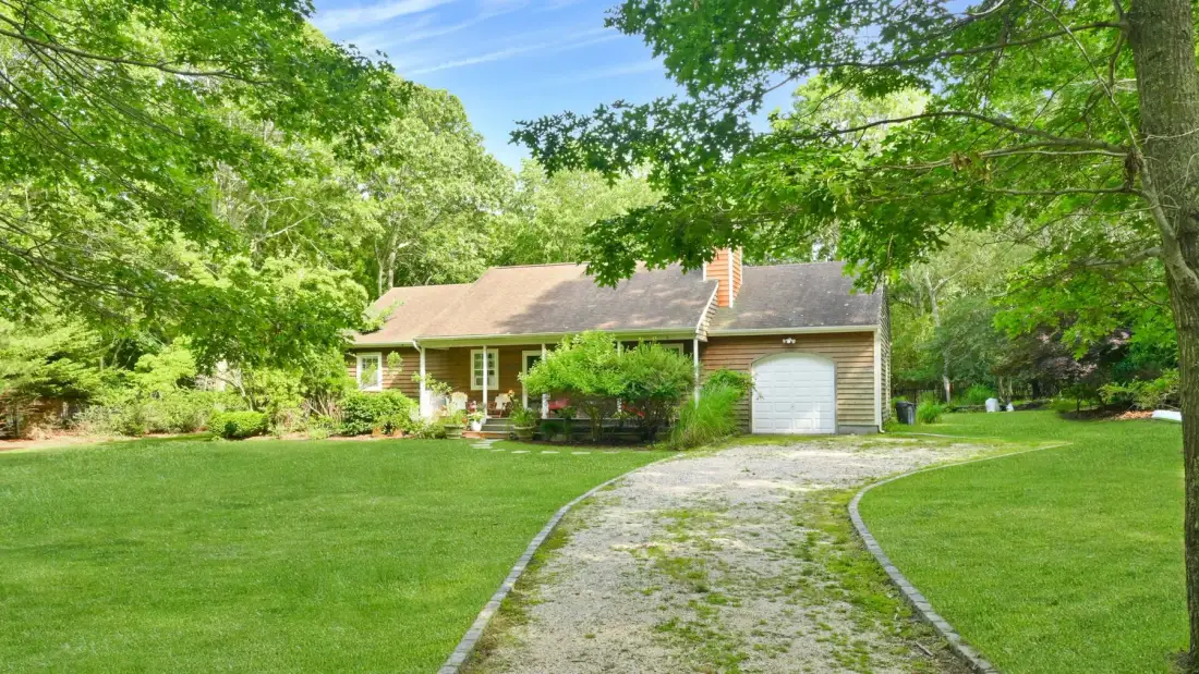 A house sits beside a curved driveway, surrounded by green trees and lawn.