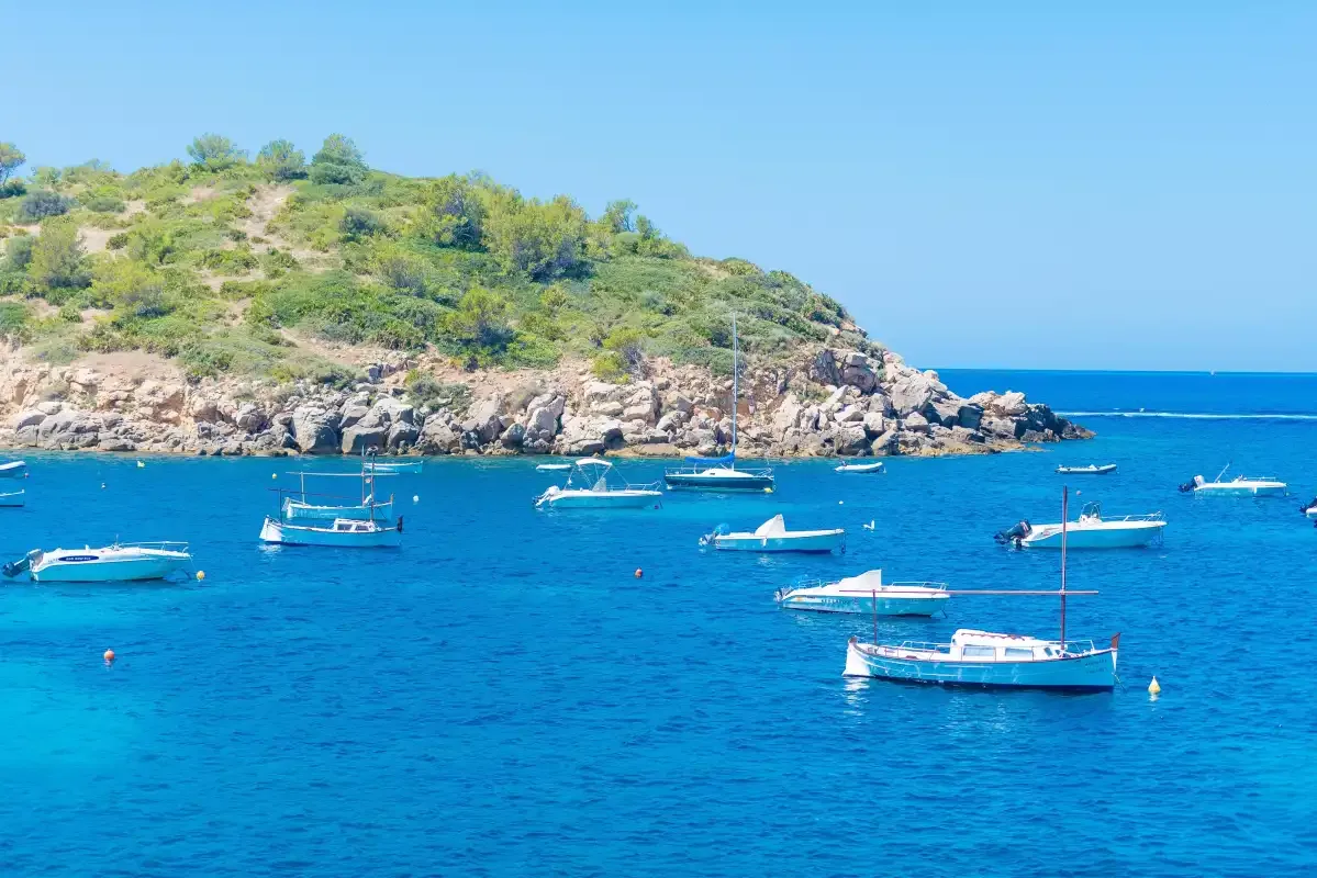 Boats float in a blue bay beside a rocky, shrub-covered headland.