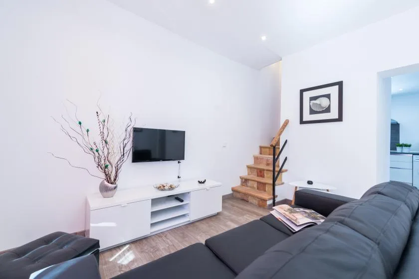 A bright living room with dark seating, a wall-mounted television, and a small staircase.