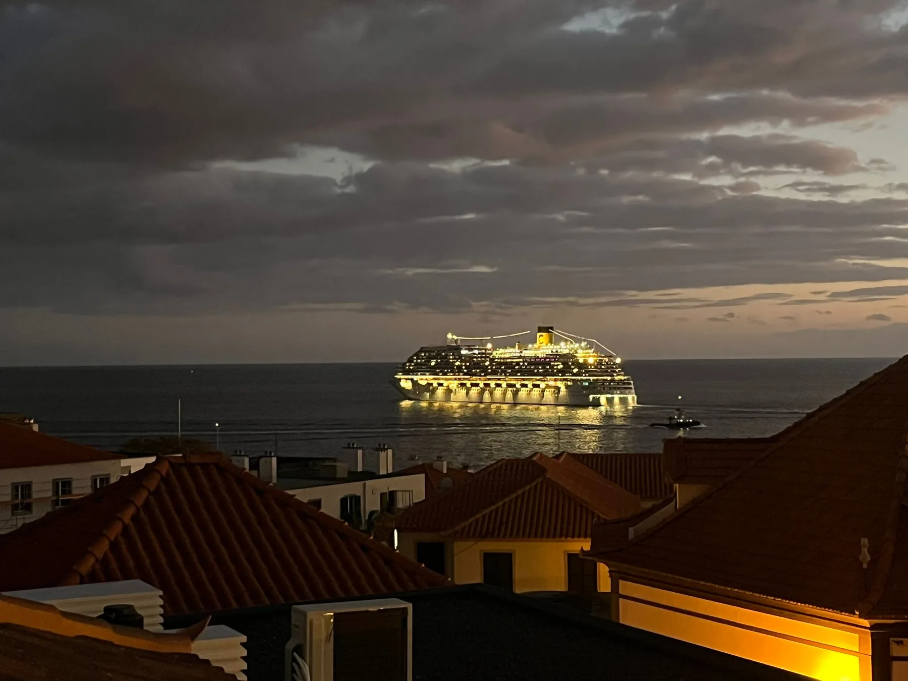 Un crucero iluminado está mar adentro, más allá de los tejados al anochecer.