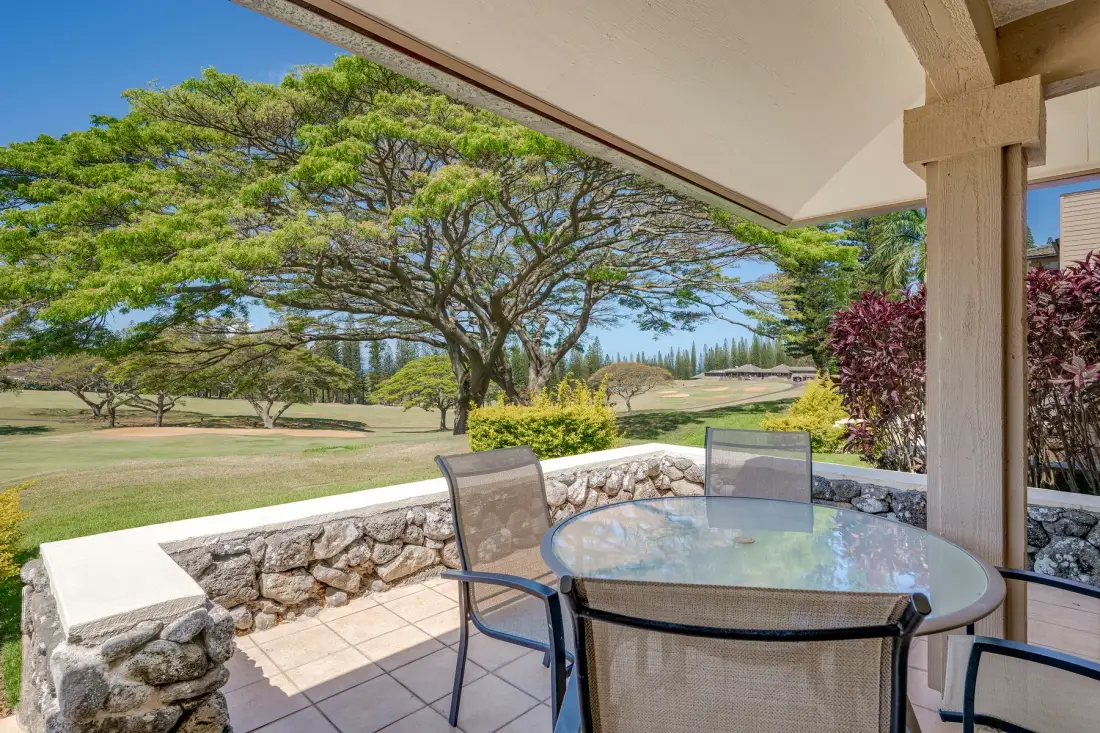 Un patio cubierto tiene una mesa de vidrio con vistas a un césped ajardinado y árboles.