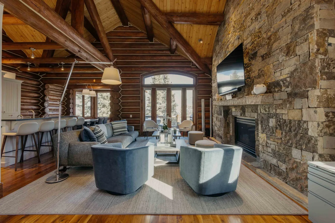 A log-walled living room with seating arranged around a coffee table and stone fireplace.