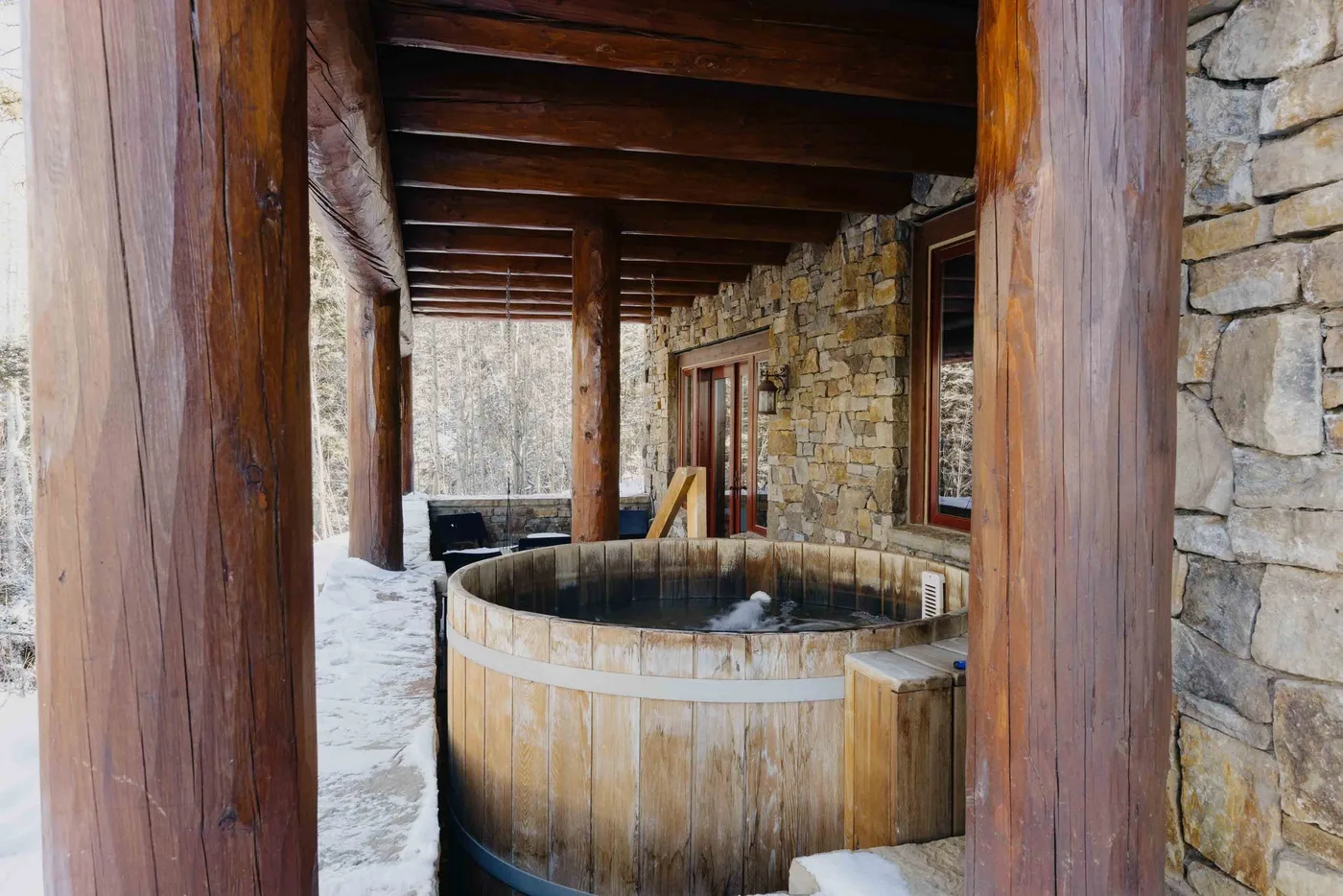 A wooden hot tub sits beneath a timber-covered outdoor area beside stone walls.