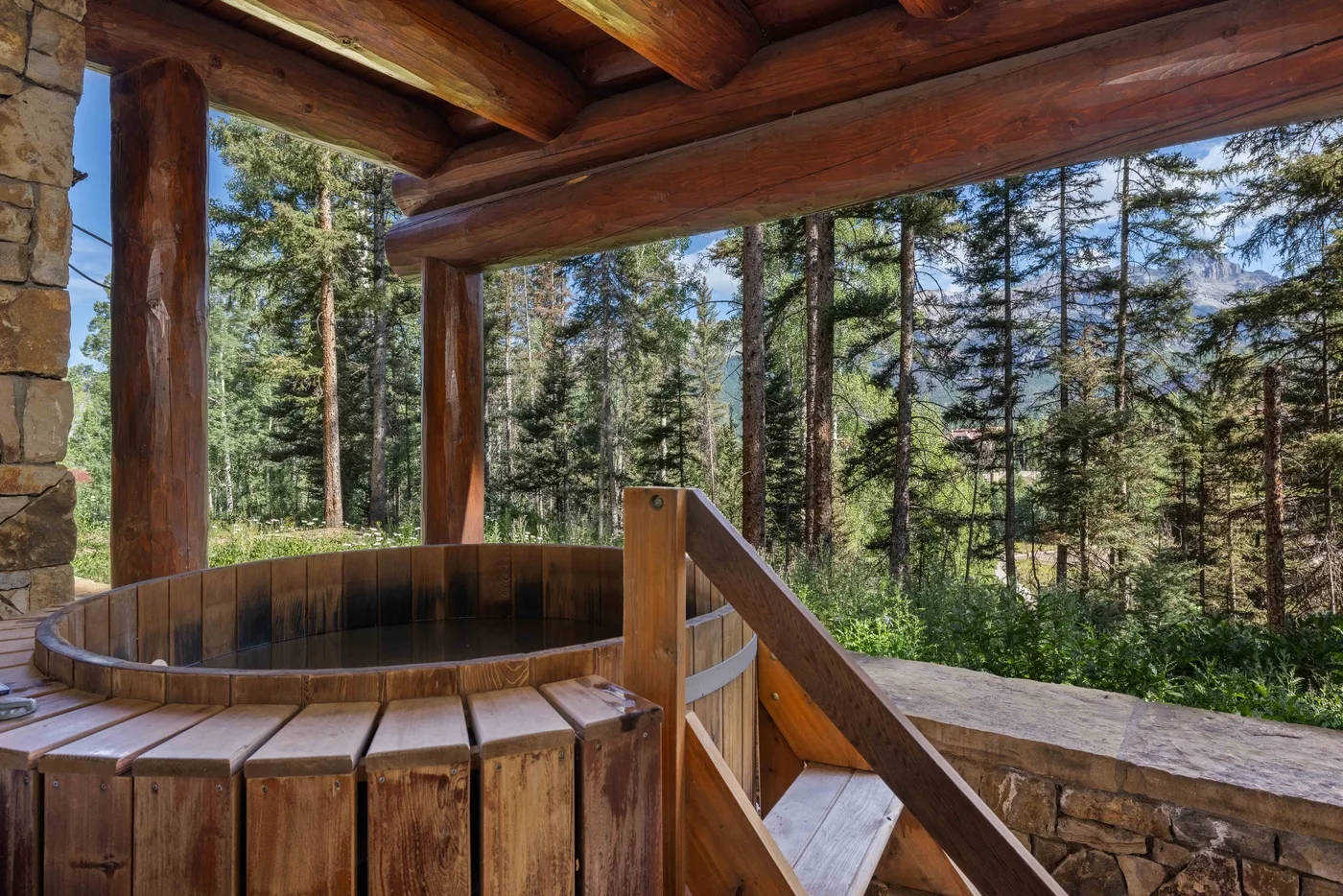 A wooden hot tub sits on a covered deck overlooking a forest.