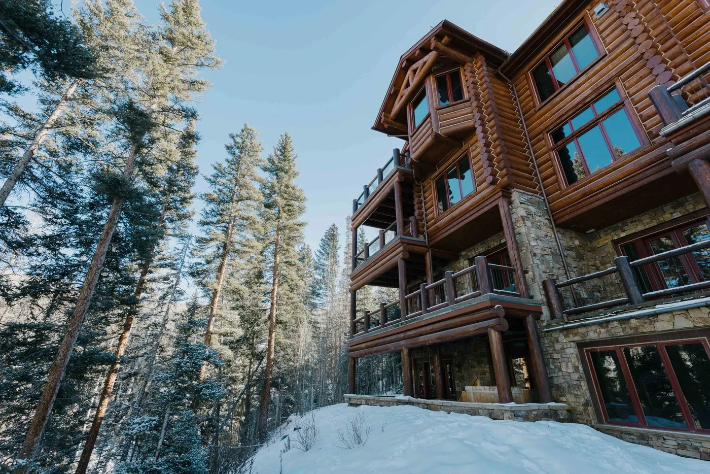 A large log building stands beside snow-covered ground and tall evergreen trees.