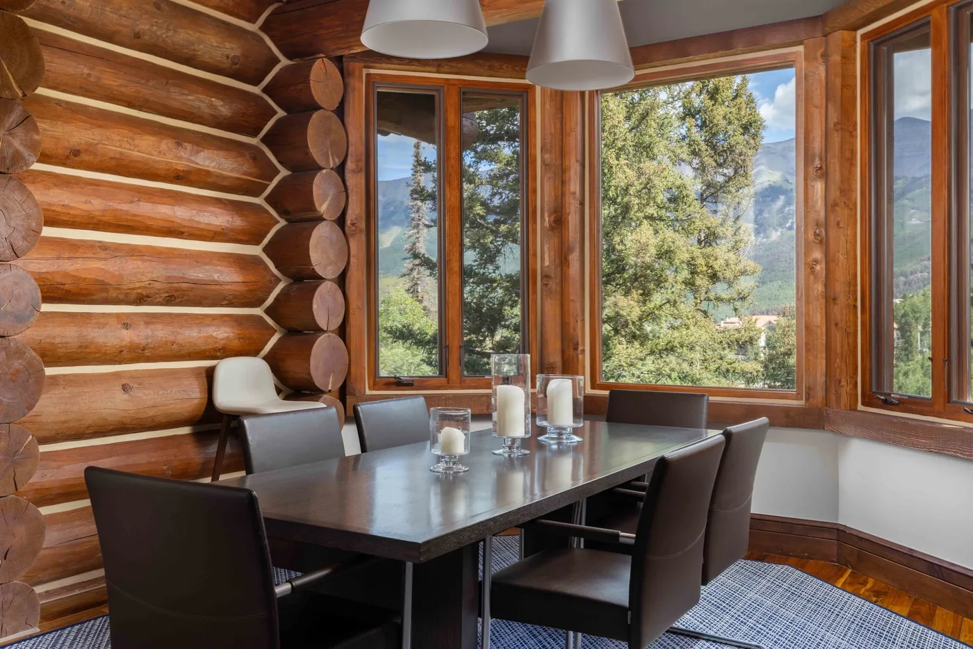 A dining area with a table and seating beside large windows overlooking trees.