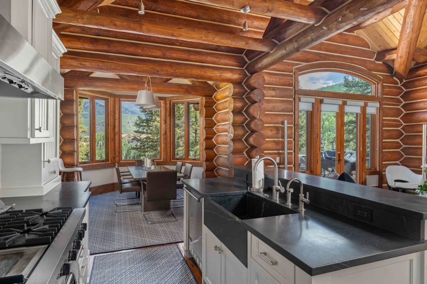 A log-cabin kitchen opens onto a dining area beside large windows.