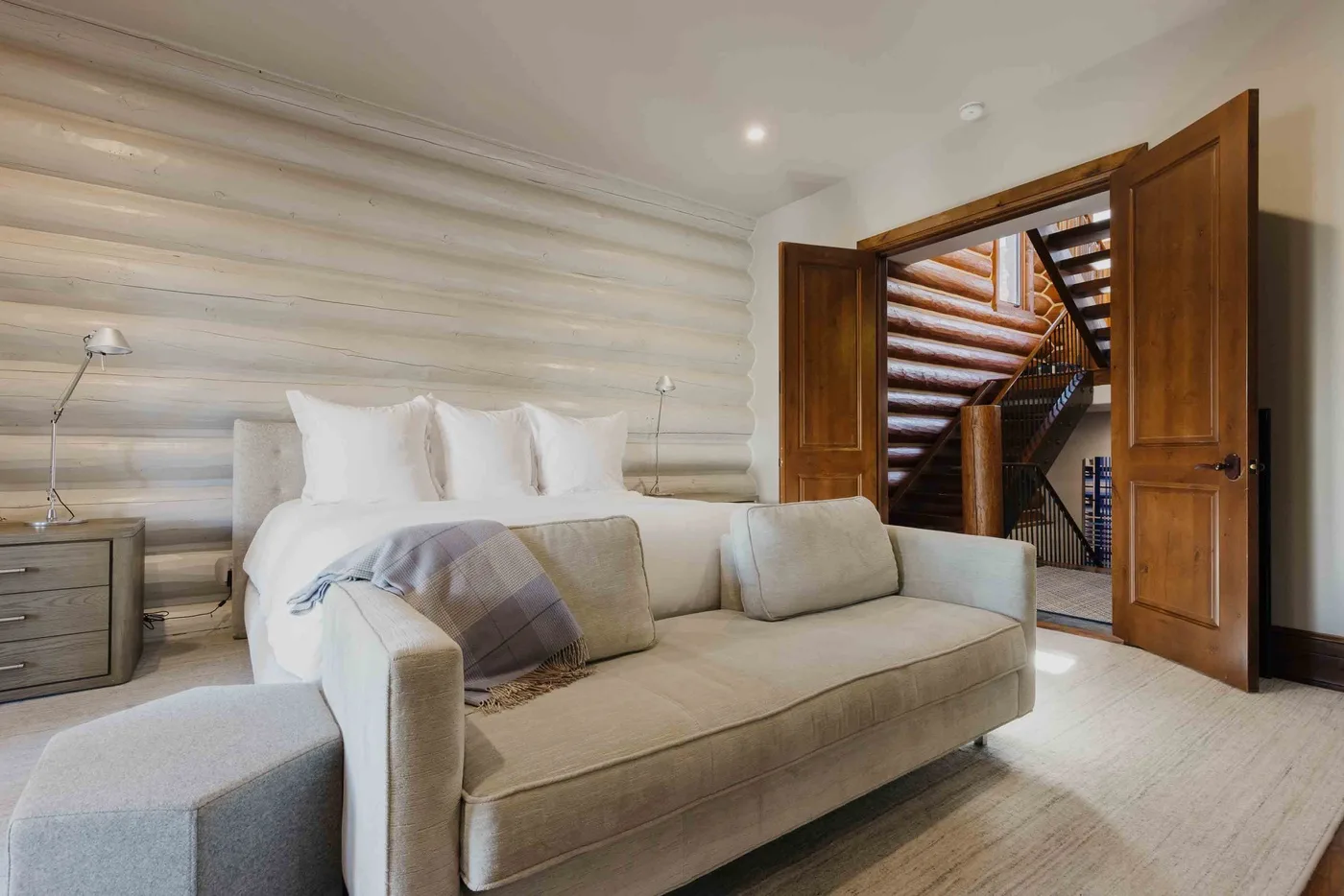A bedroom with a bed behind a sofa and open wooden doors.
