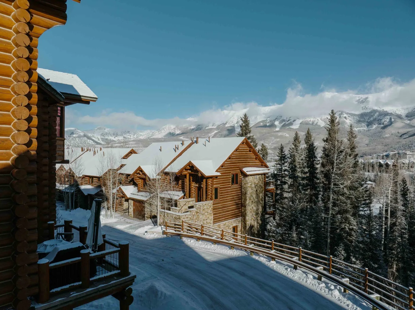 Snow-covered log cabins sit beside a wooded slope, with mountains in the distance.