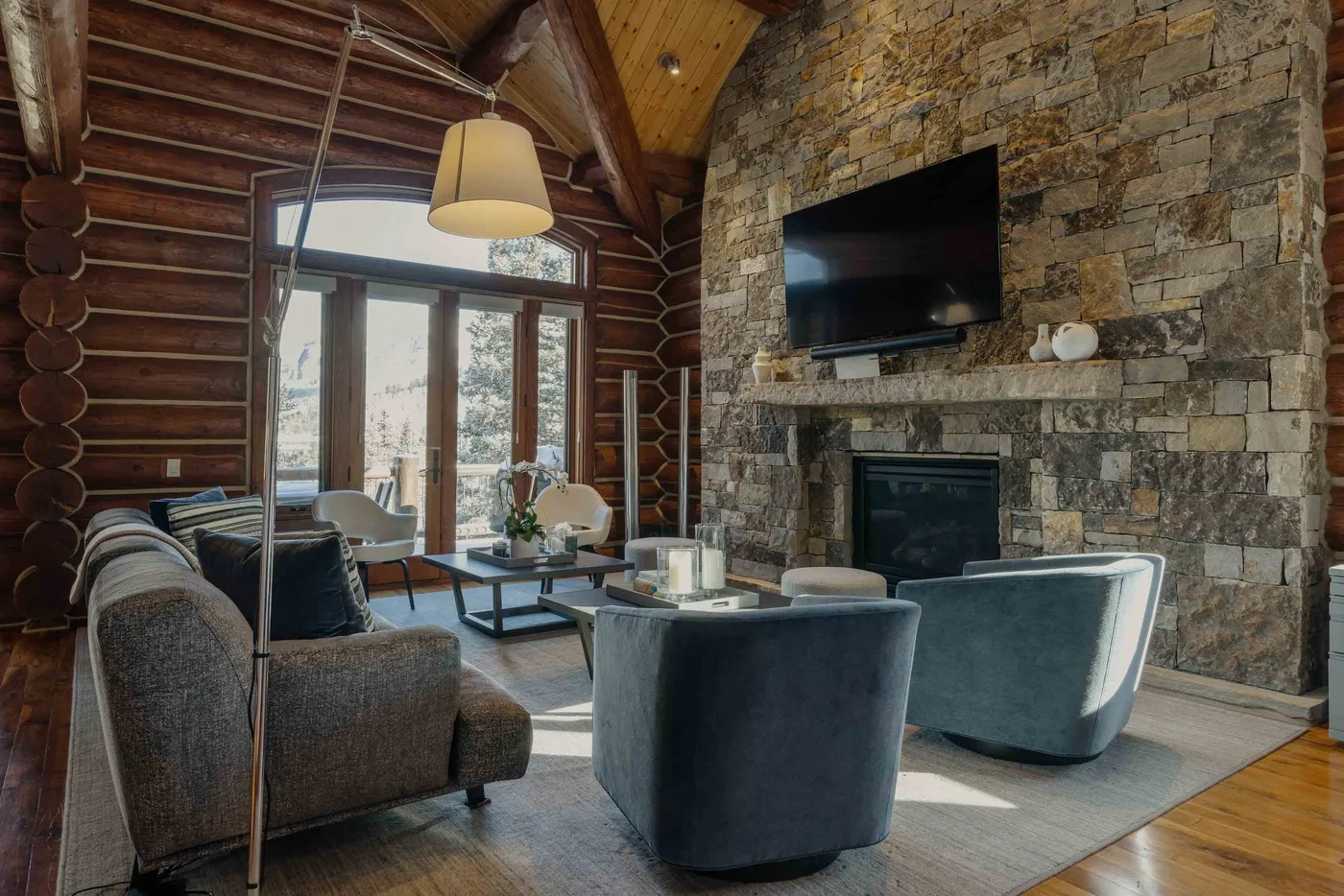 A log cabin living room with seating arranged around a stone fireplace and television.