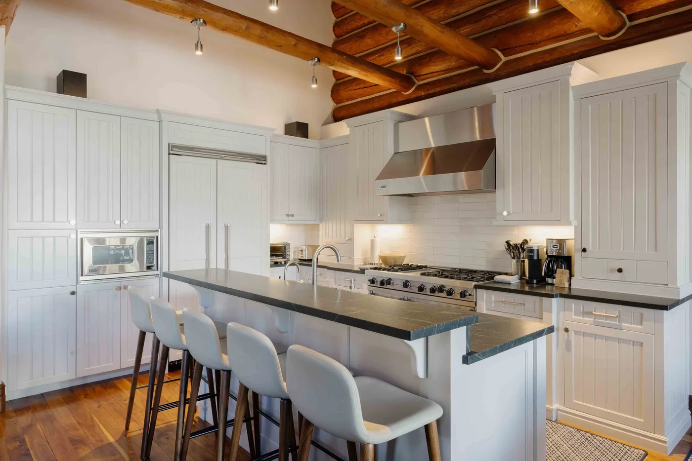 A bright kitchen with a long island, pale cabinetry, and exposed wooden ceiling beams.