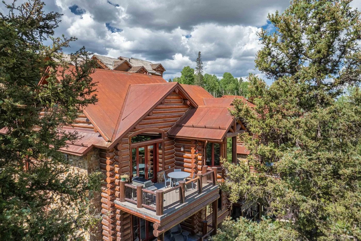 A log cabin with red roof sections and a small balcony, surrounded by trees.
