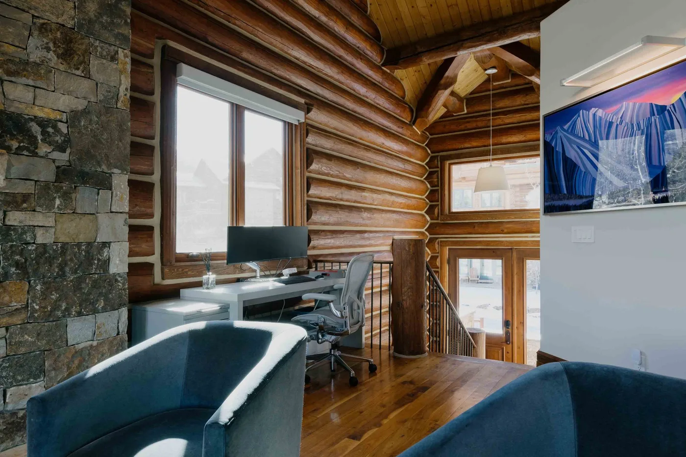 A log-cabin office with a desk beside a large window and a stair railing.