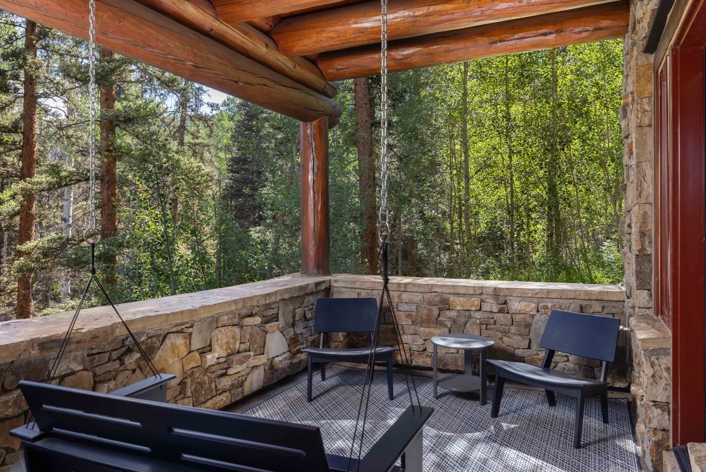 A covered stone patio with outdoor seating overlooks a wooded area.