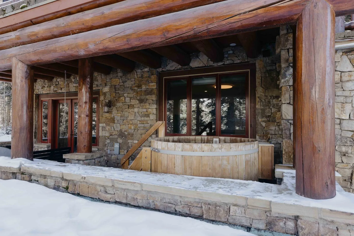 A wooden hot tub sits beneath a timber-covered patio against a stone wall.