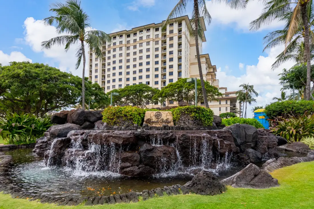 Una cascada ornamental se encuentra frente a un alto edificio hotelero rodeado de vegetación tropical.