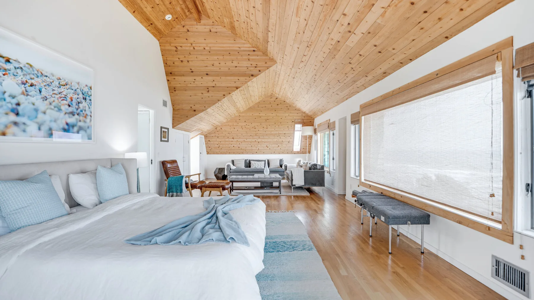 A bright bedroom opens into a sitting area beneath a vaulted wood ceiling.