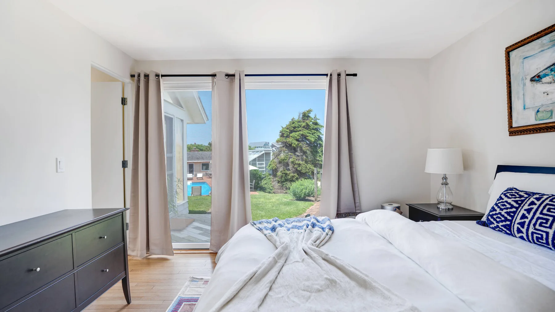 A bright bedroom with a bed, bedside table, and large windows overlooking greenery.