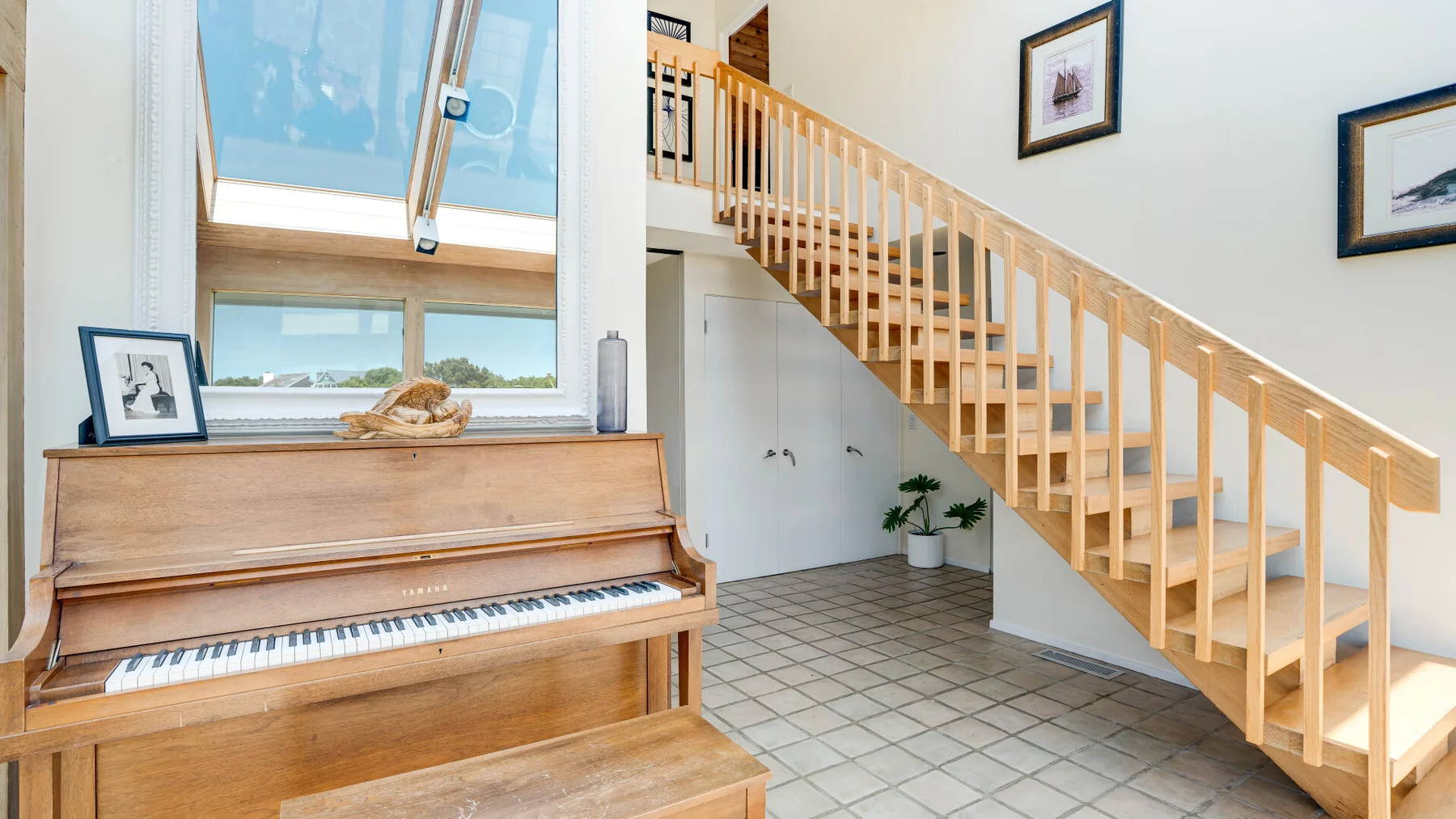 A bright room features a wooden piano beneath a large window and a staircase.