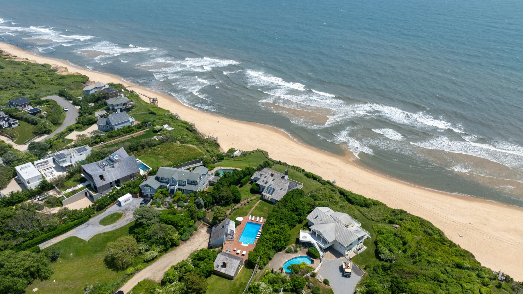 An aerial view shows homes among green vegetation beside a sandy beach and ocean.